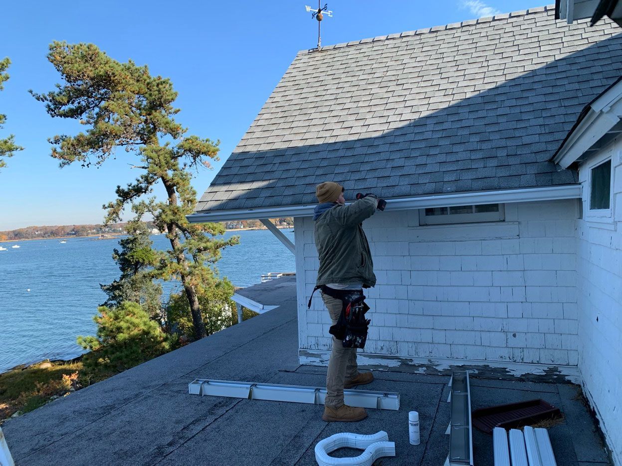A person in work gear installs gutters on the white brick wall of a coastal home overlooking a blue bay.