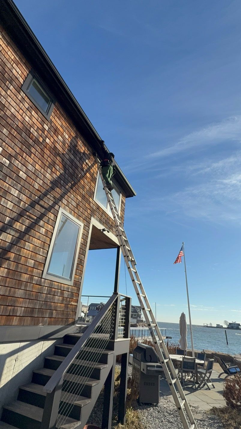 A person on a tall ladder reaches toward the roof edge of a brick building near a calm body of water under a blue sky.