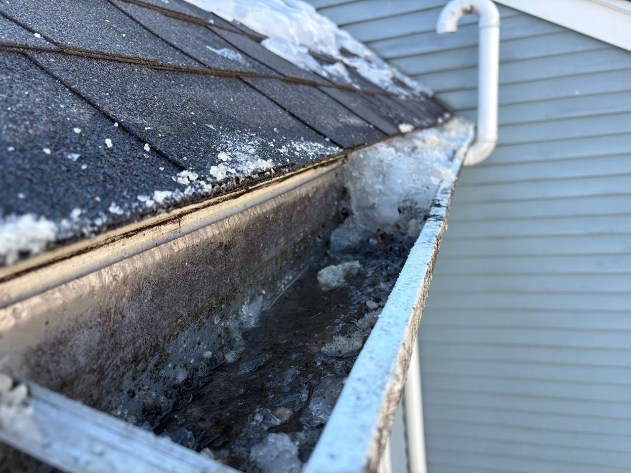 A close-up view of a metal roof gutter filled with melting snow and ice, attached to the side of a grey-sided house.