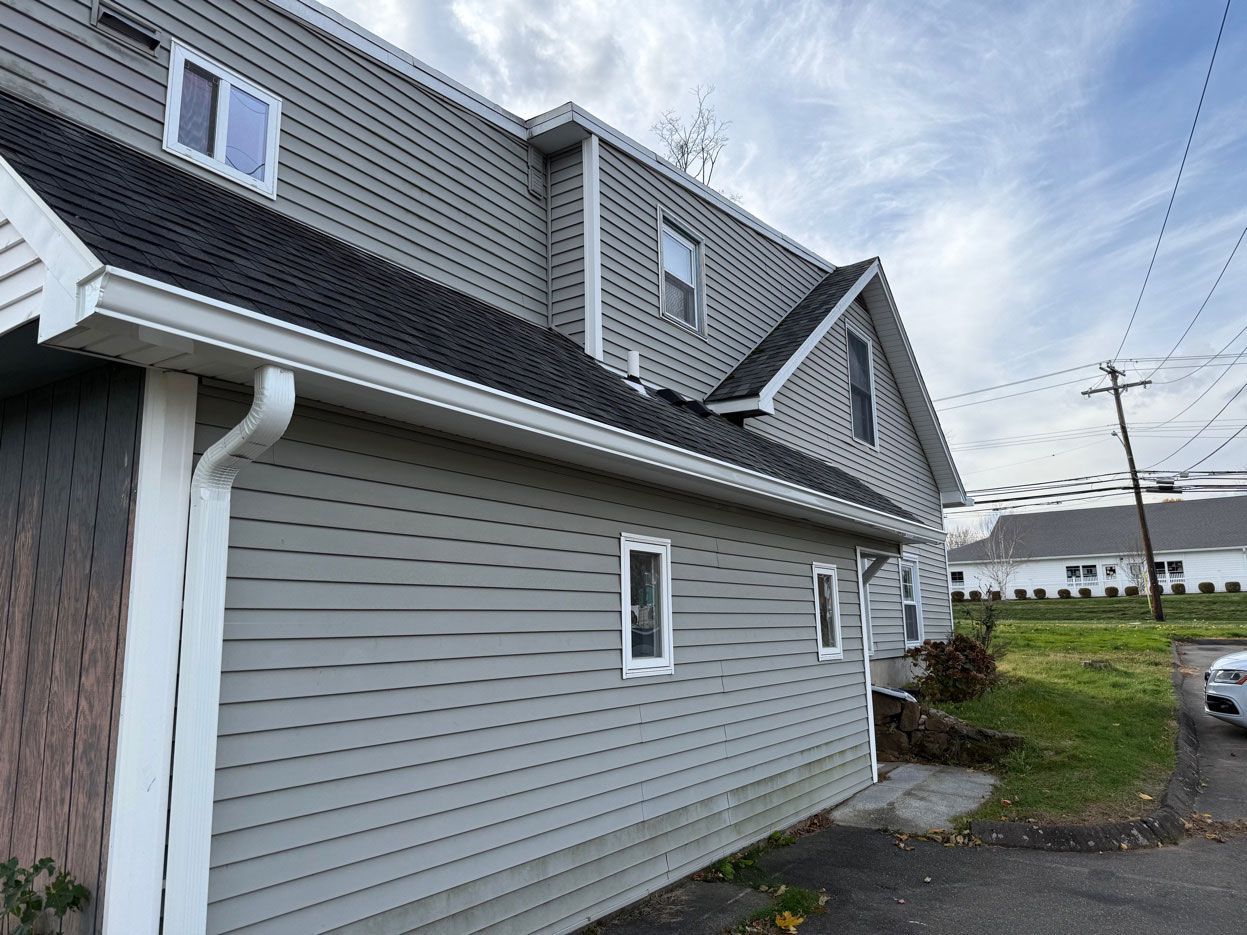 A two-story building with gray vinyl siding, black shingled roofs, and several windows under a partly cloudy sky.