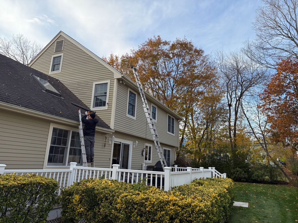 A person climbs an extension ladder leaned against the tan siding of a two-story house with a white deck in autumn.