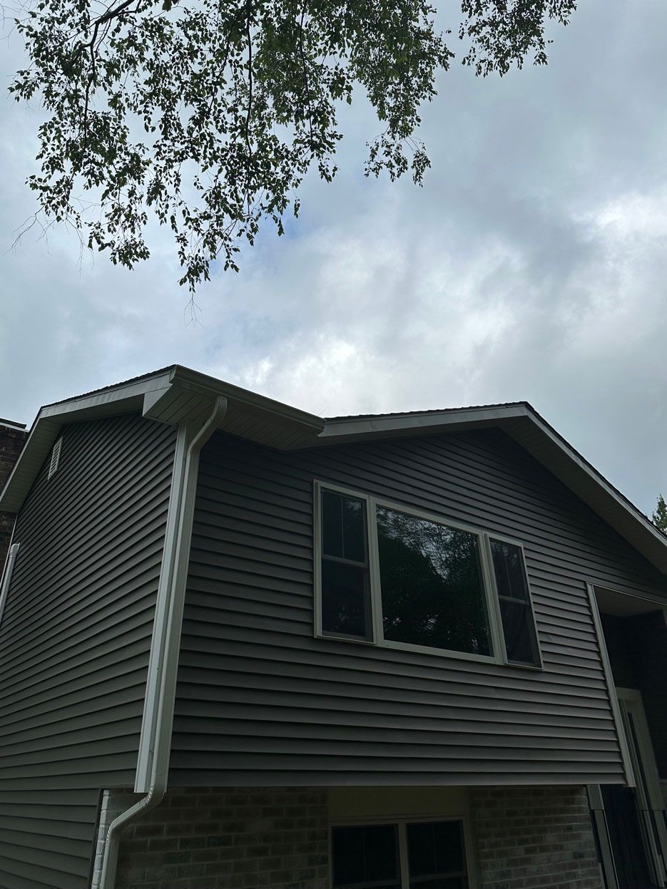 A low-angle view of a grey-sided house with a white gutter system and a brick foundation under a cloudy sky.