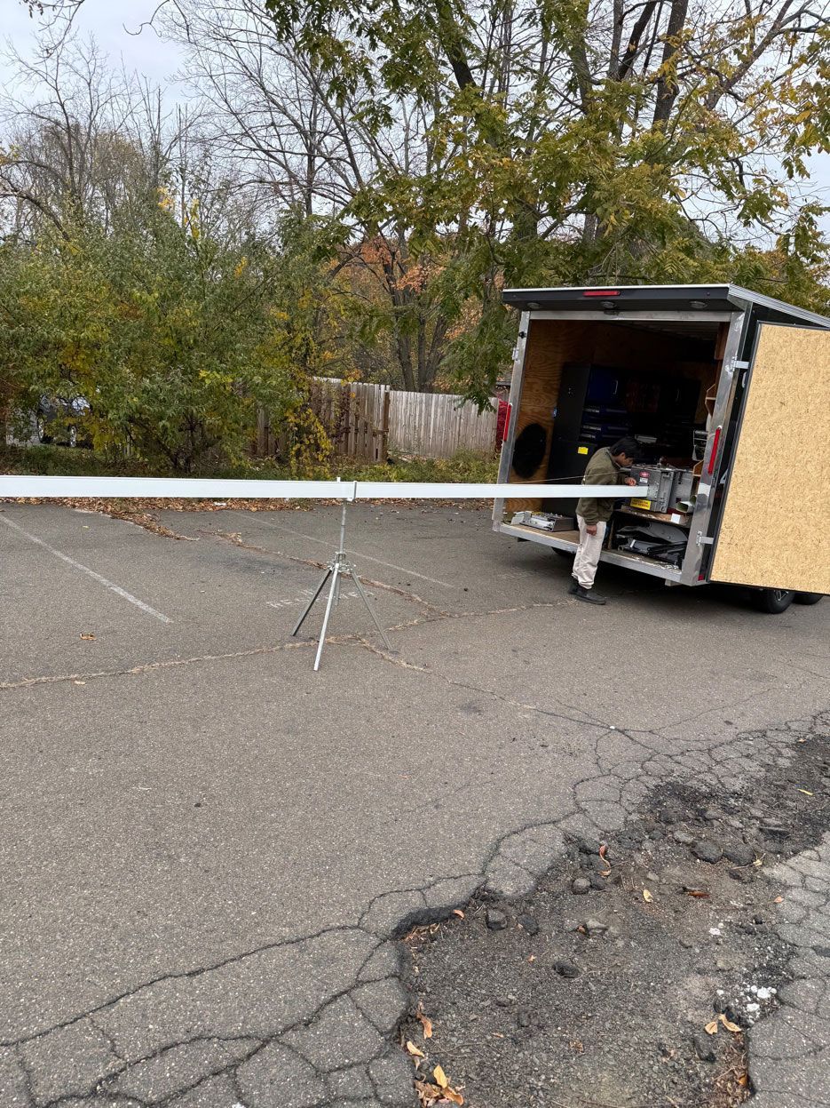 A person works at the rear of an open utility trailer, supporting a long metal beam on a tripod in an outdoor lot.