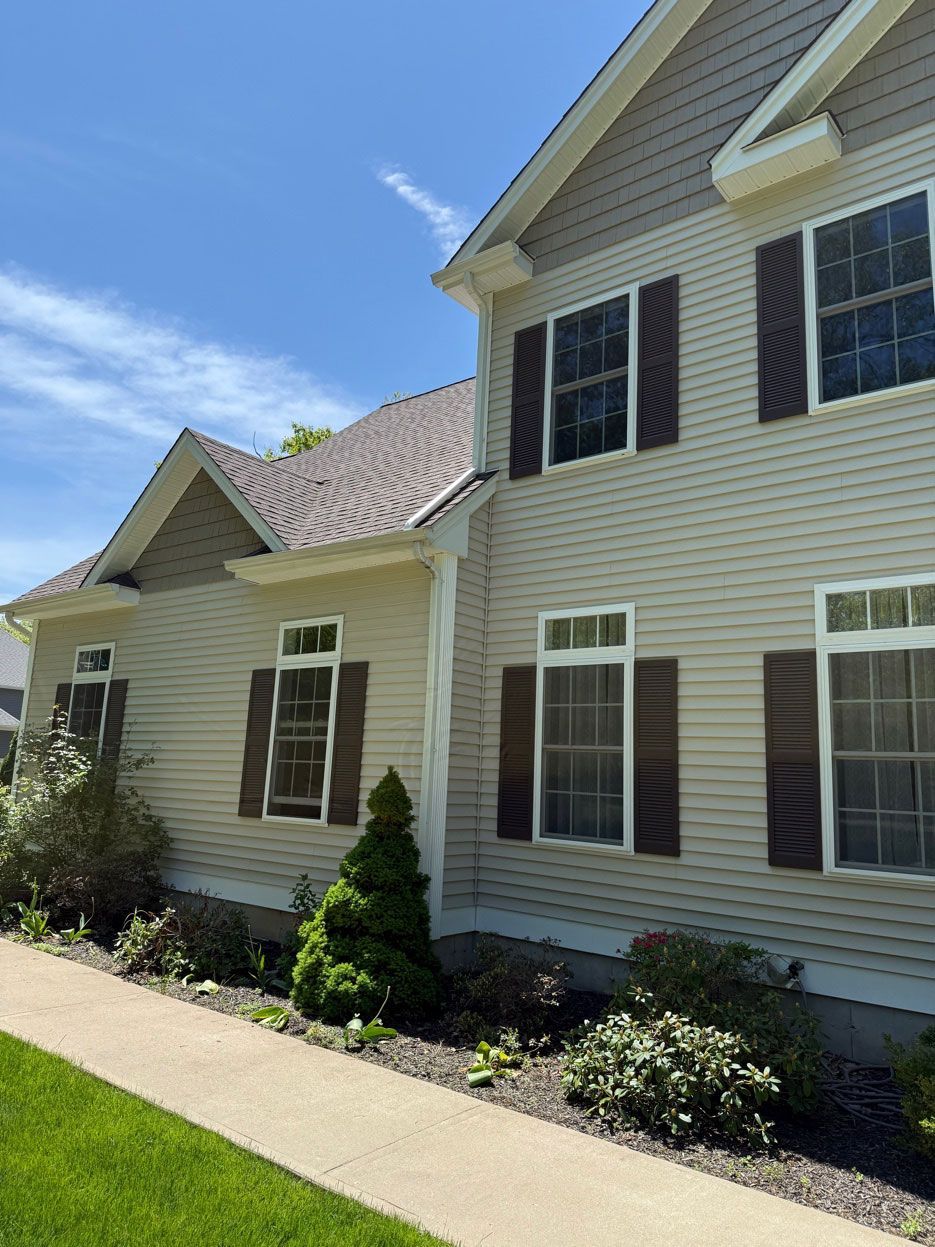 A light-colored two-story house with dark shutters, a small gabled roof section, and a manicured lawn under a blue sky.