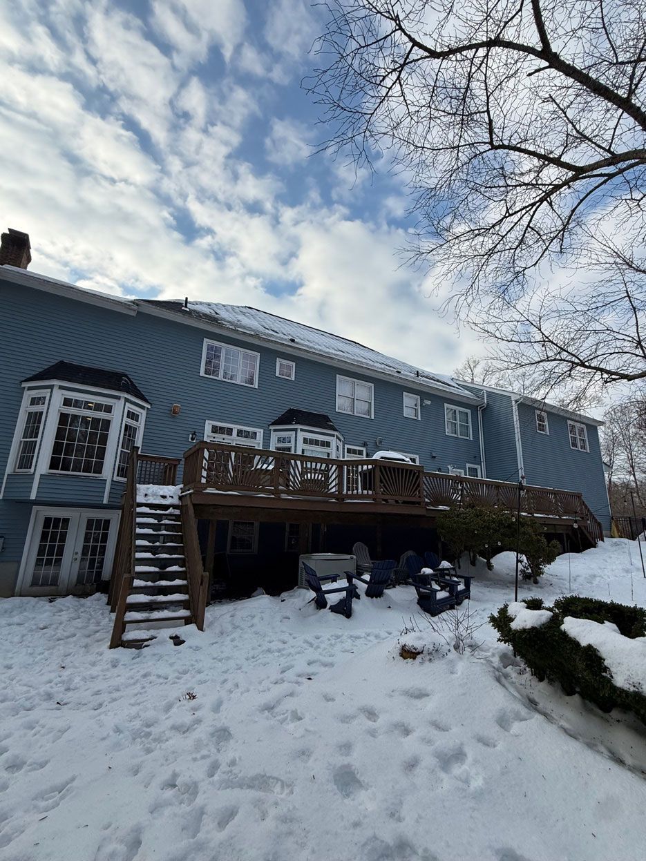 Blue house with a wooden deck and staircase, surrounded by snow under a partly cloudy sky.