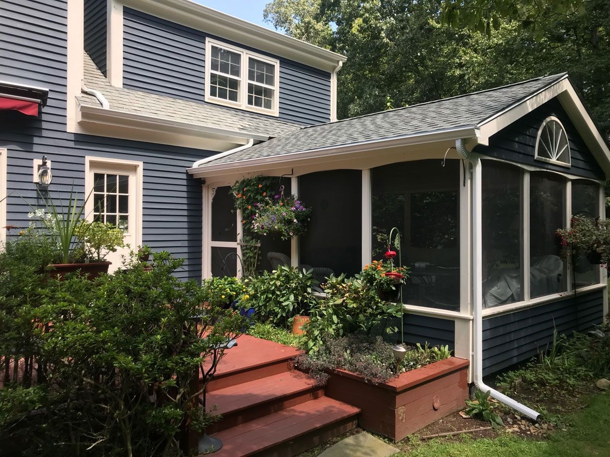 A blue-sided two-story house with a screened-in porch, wooden steps, and surrounding garden greenery.