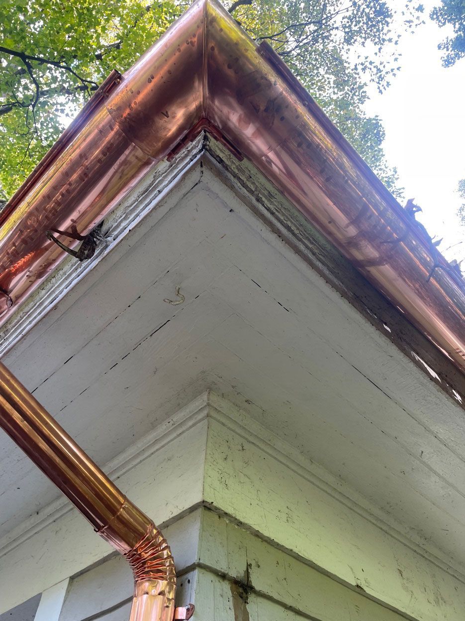 Low-angle shot of a copper gutter system installed on the white eave of a house corner, with a downspout attached.
