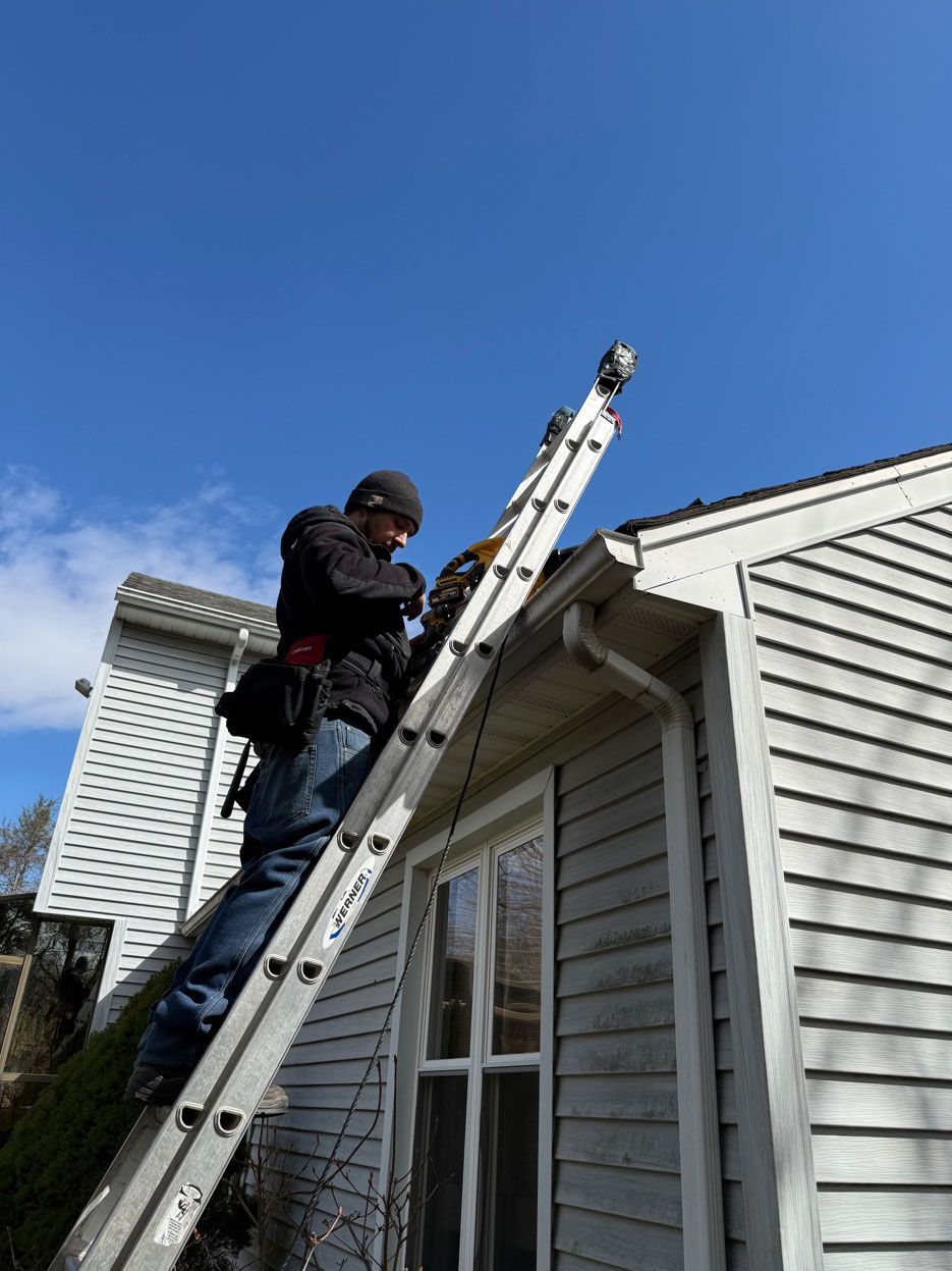 A person in a black jacket and beanie climbing an extension ladder against the side of a light-colored house.