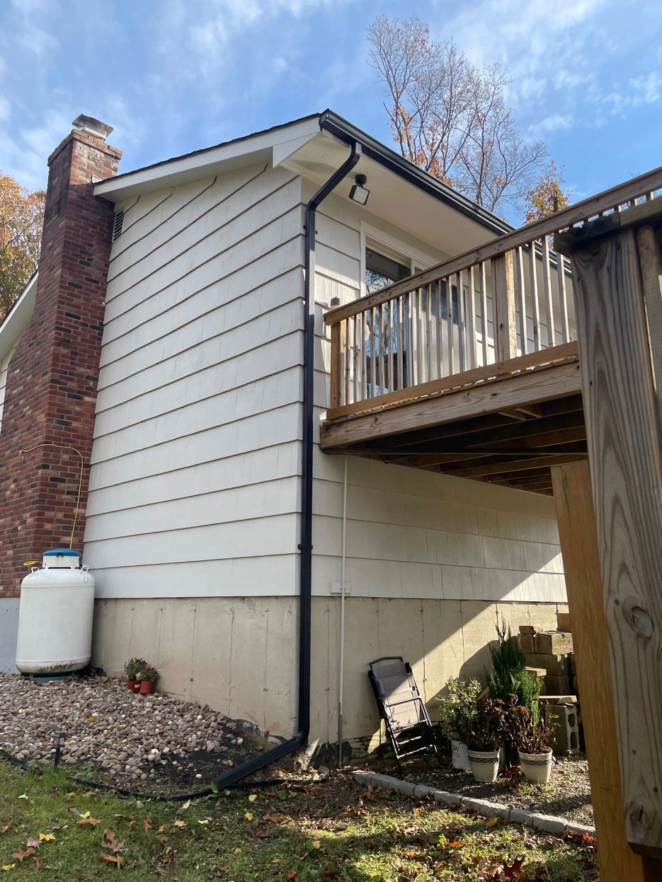 A side view of a white-sided house with a brick chimney, a raised wooden deck, a downspout, and a white tank on gravel.