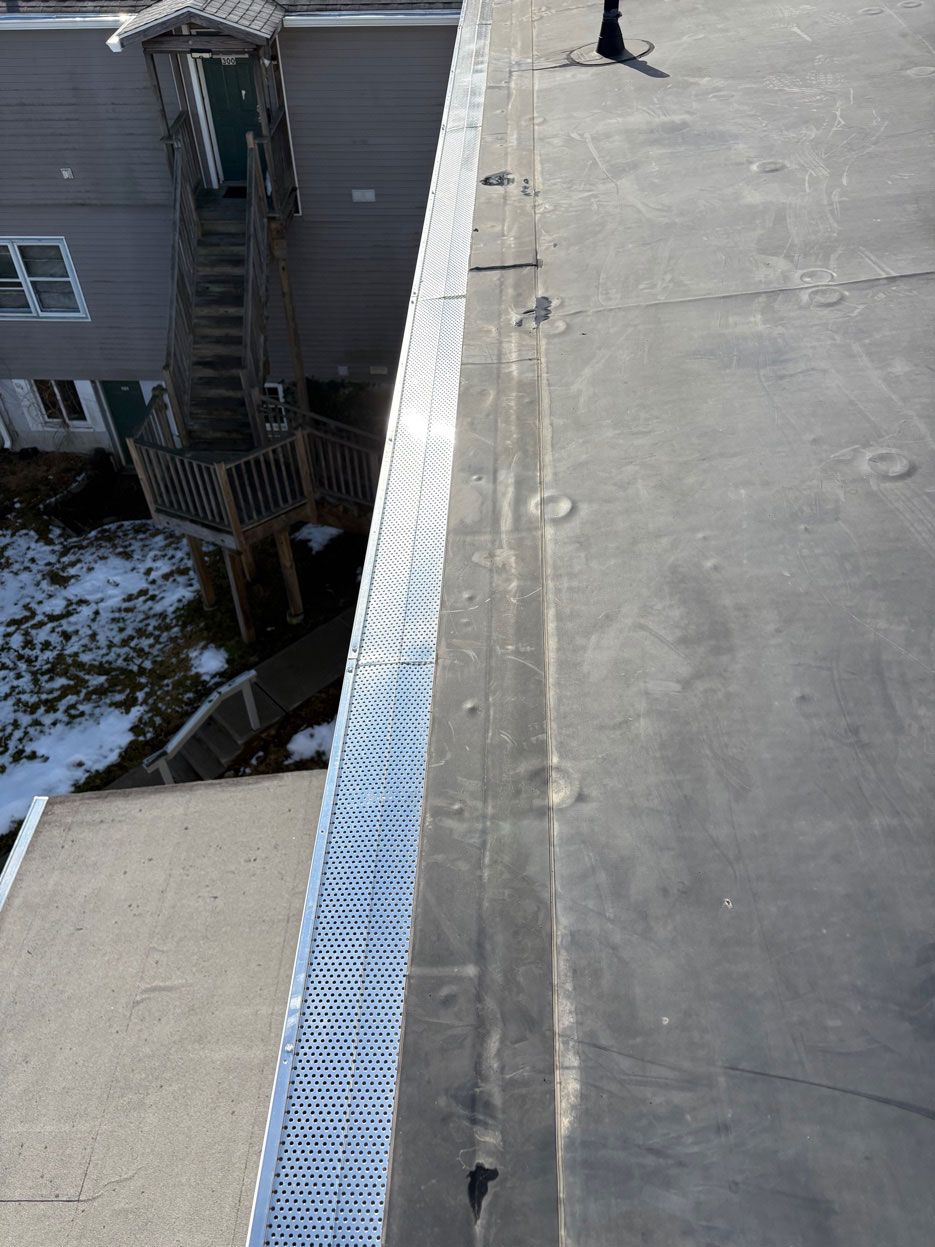 A high-angle view of a flat, gray roof edge with a perforated metal gutter guard, overlooking a building and snowy ground.