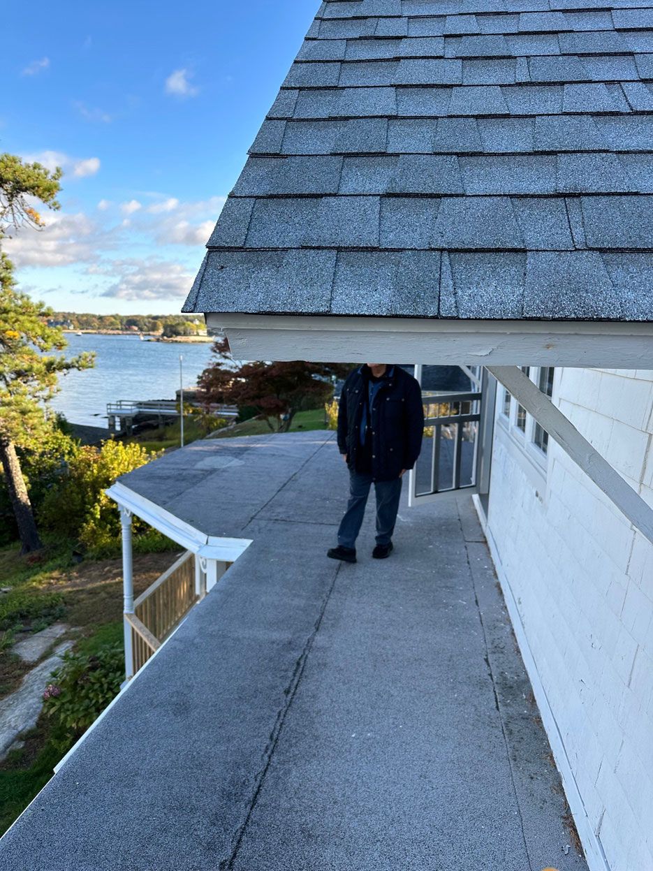 A person stands on a flat balcony overlooking a body of water, positioned beneath the low overhang of a shingled roof.