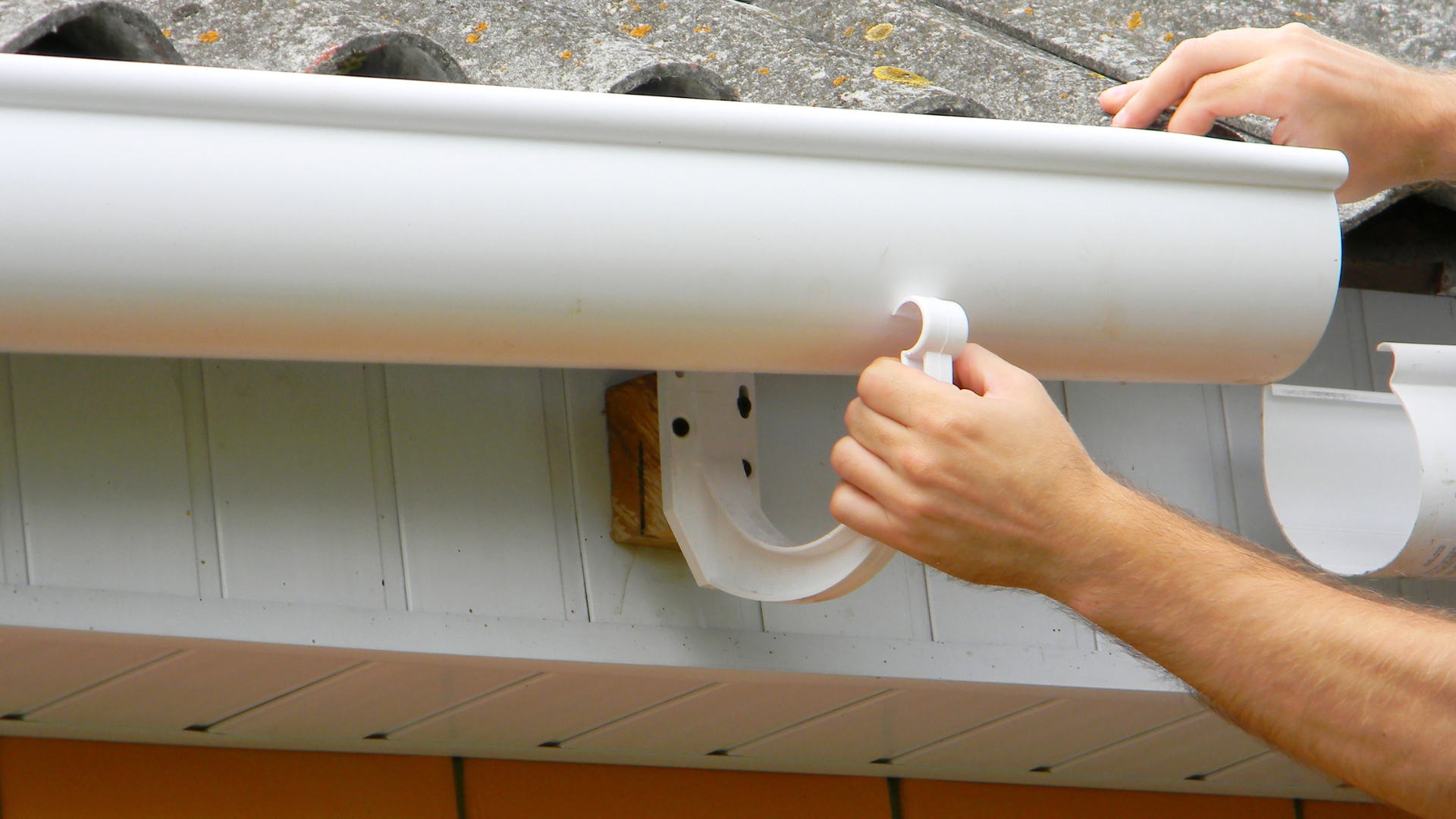 Person installing a white gutter on a house roof eave with a hand tool.