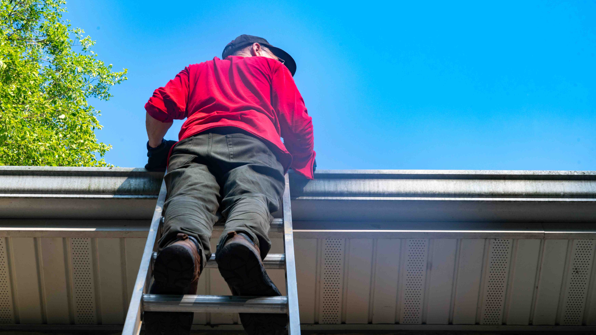 Person in a red jacket on a ladder cleaning a roof gutter against a blue sky