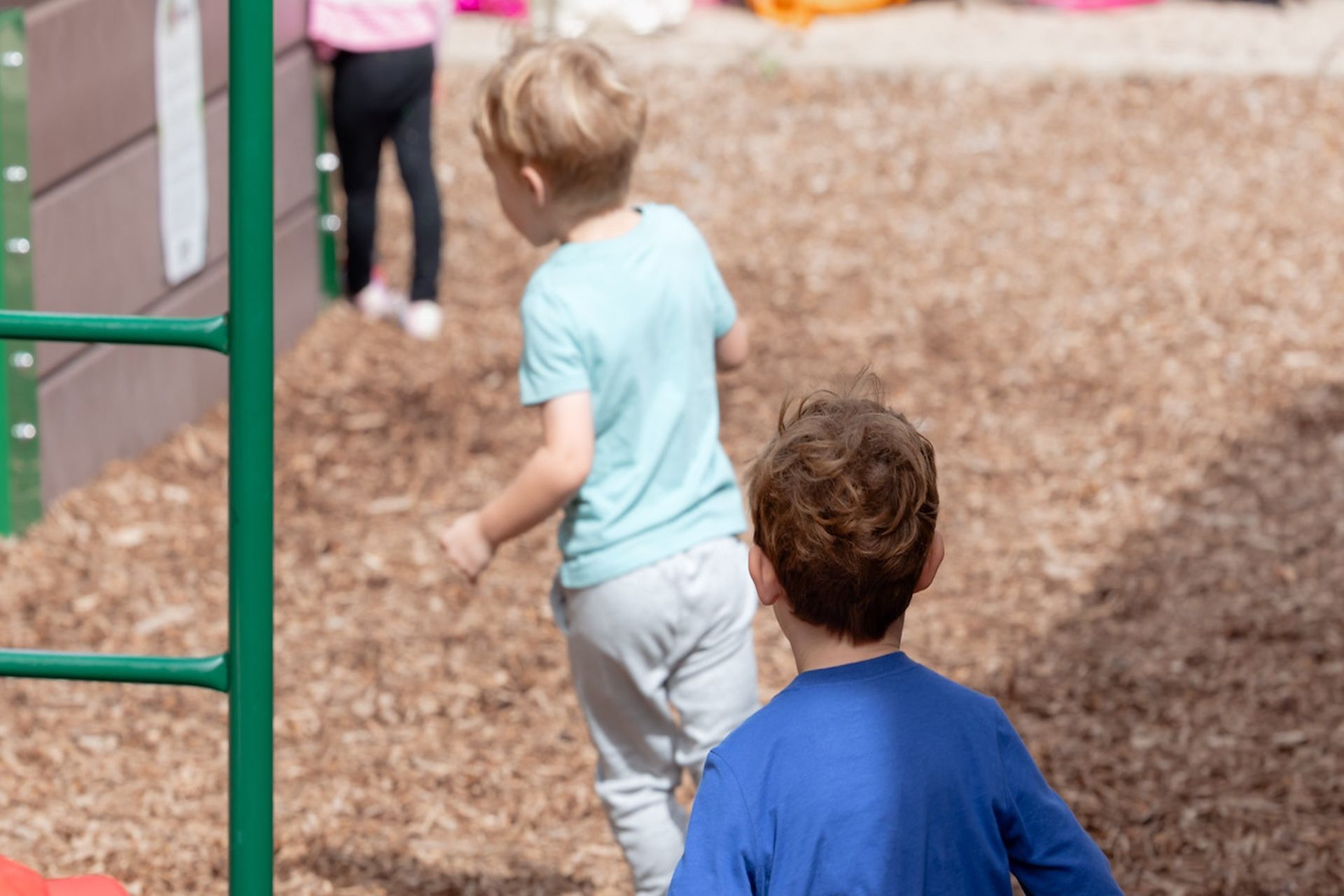 Children play on a wood-chip covered playground near a green metal structure.