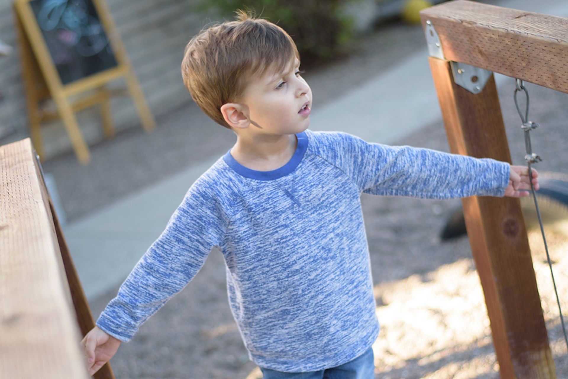 A child in a blue heathered long-sleeved shirt stands outdoors, reaching out toward a wooden playground structure.