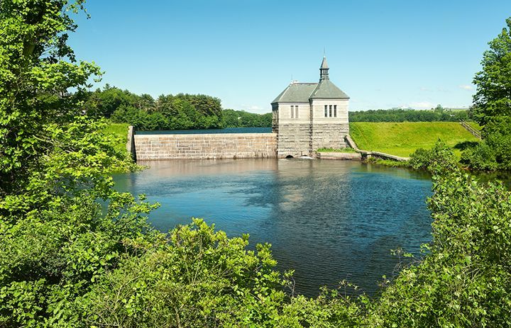 Small stone building beside a calm blue reservoir, framed by lush green trees under a clear sky