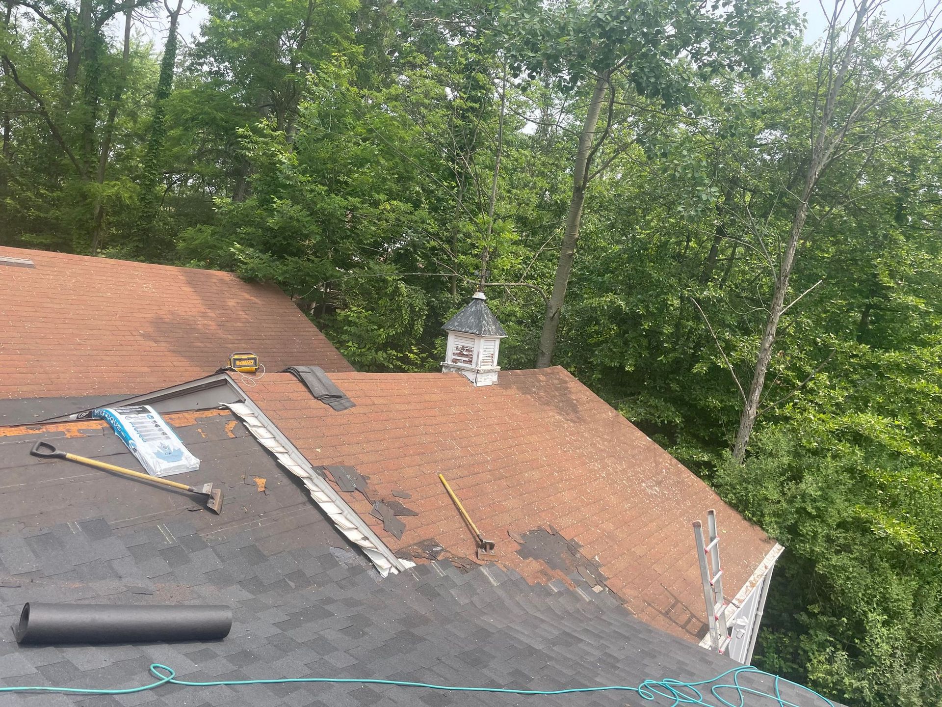 Roof with shingles being replaced; tools and materials are visible. Trees are in the background.
