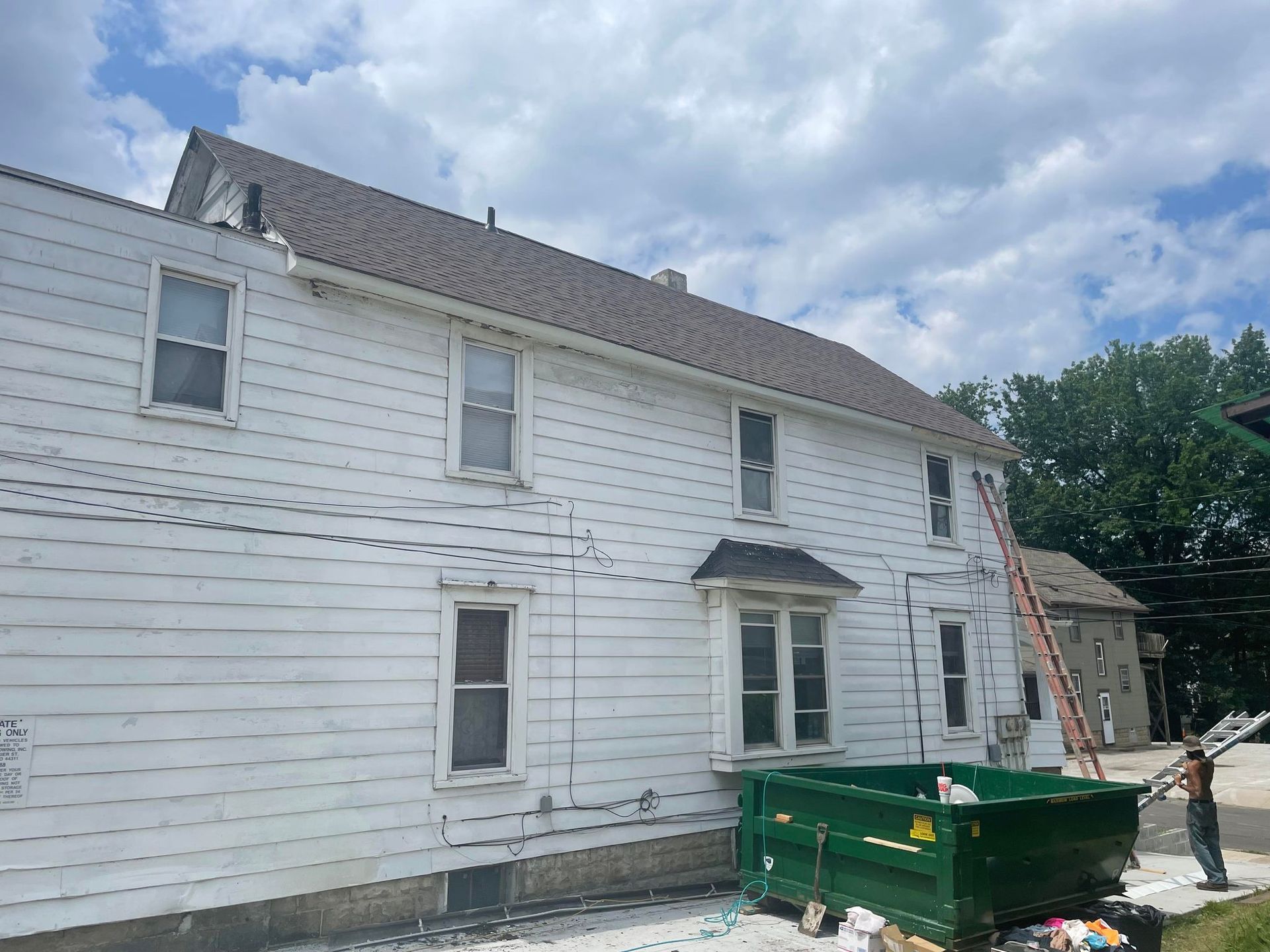 White two-story house with a brown roof. A green dumpster sits in front. Person near ladder is visible.