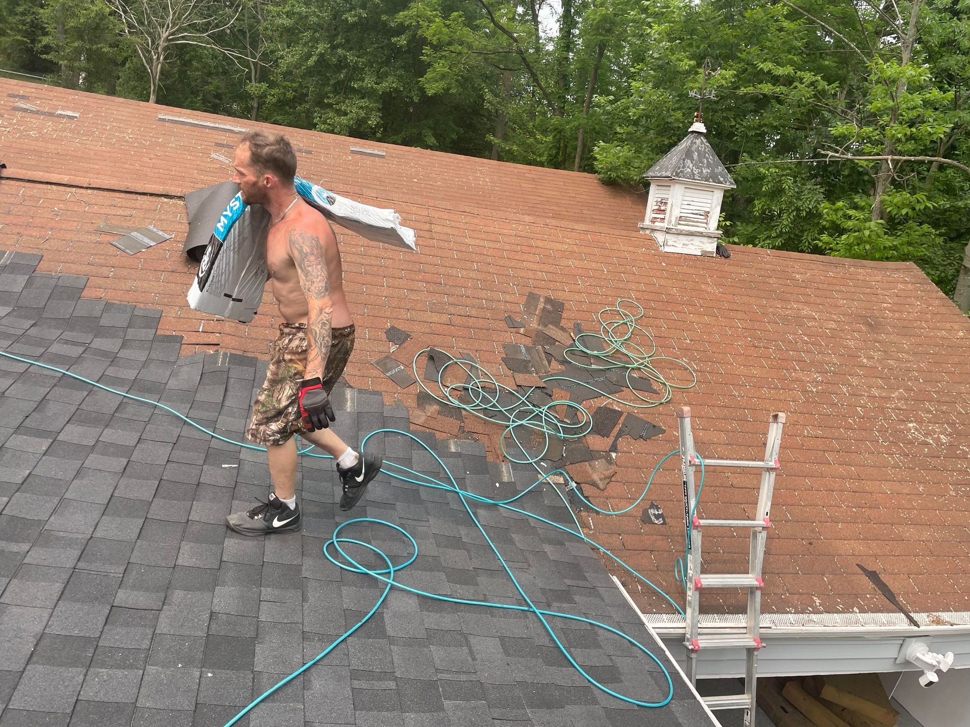 Man carrying roofing materials on a roof, near a ladder and a chimney, on a sunny day.