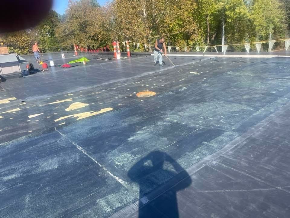 Man sweeping surface; black tarp, yellow debris, white markings. Outdoors on a sunny day.