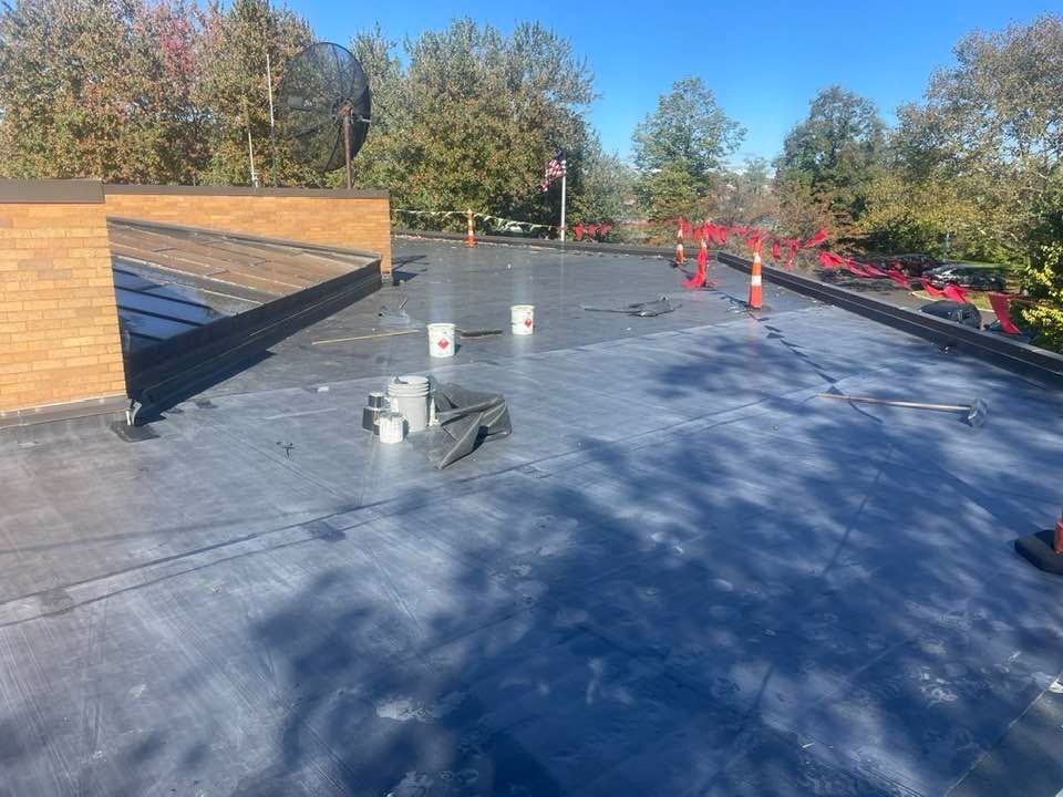 Flat roof with dark membrane, buckets, tools, and red safety equipment under a blue sky.