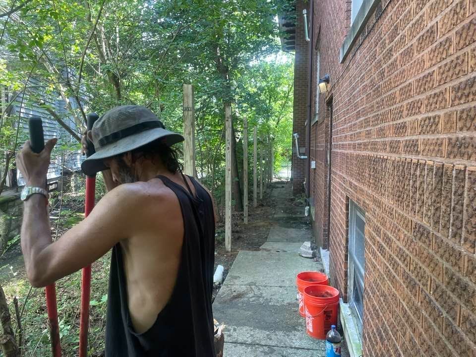 Person holding a tool, standing next to a brick building and narrow path lined with wooden posts.