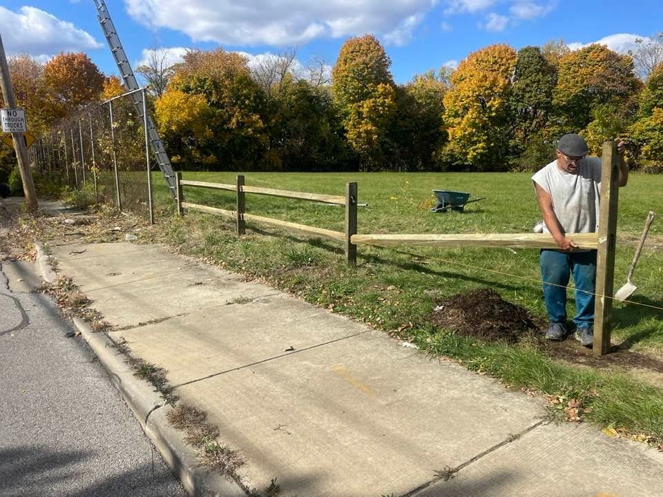 Man constructing a wooden fence next to a sidewalk and green field.