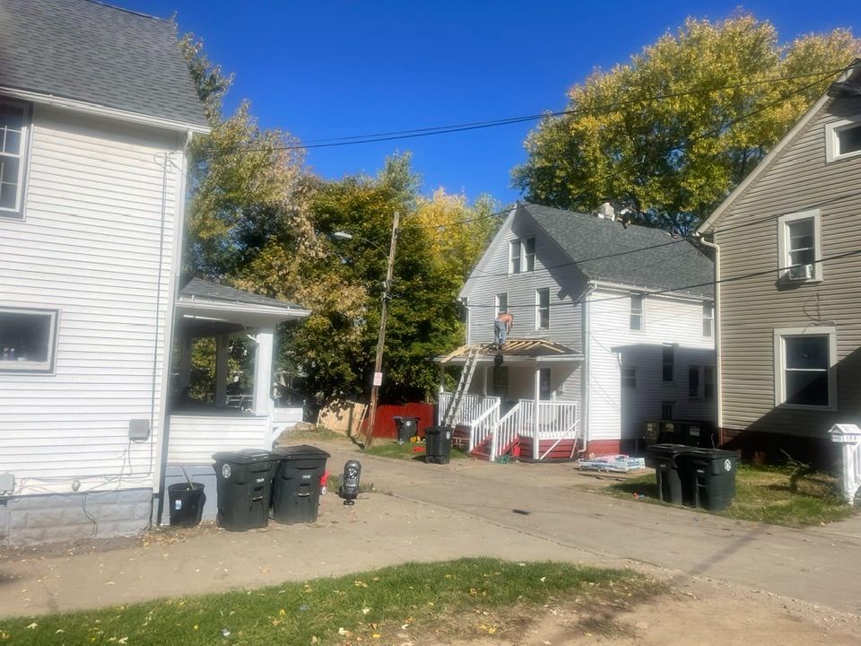 View of three houses in a yard with a driveway. One house has construction in progress, with ladders. Green and blue sky.