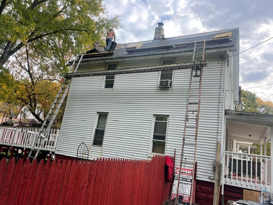 A person on a roof removing shingles, two ladders against the house, red fence in foreground.
