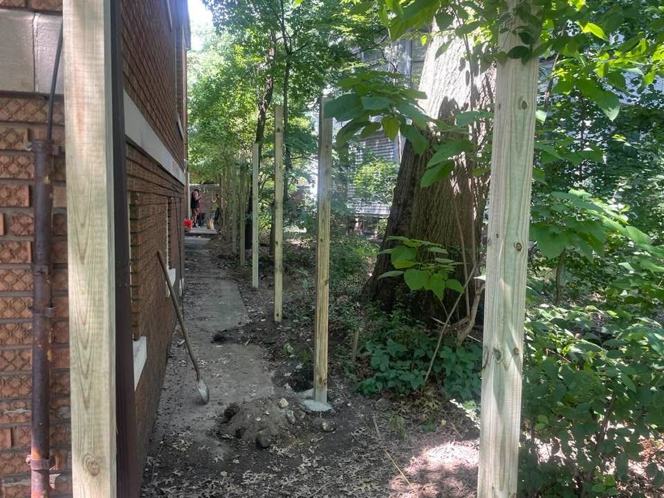 Narrow outdoor path with trees, building on one side, wooden posts and dirt.