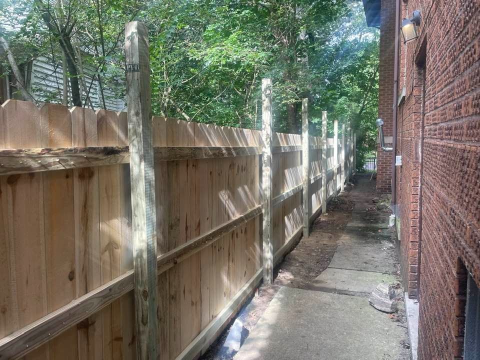 Wooden fence alongside a brick building and a narrow concrete walkway, trees in the background.