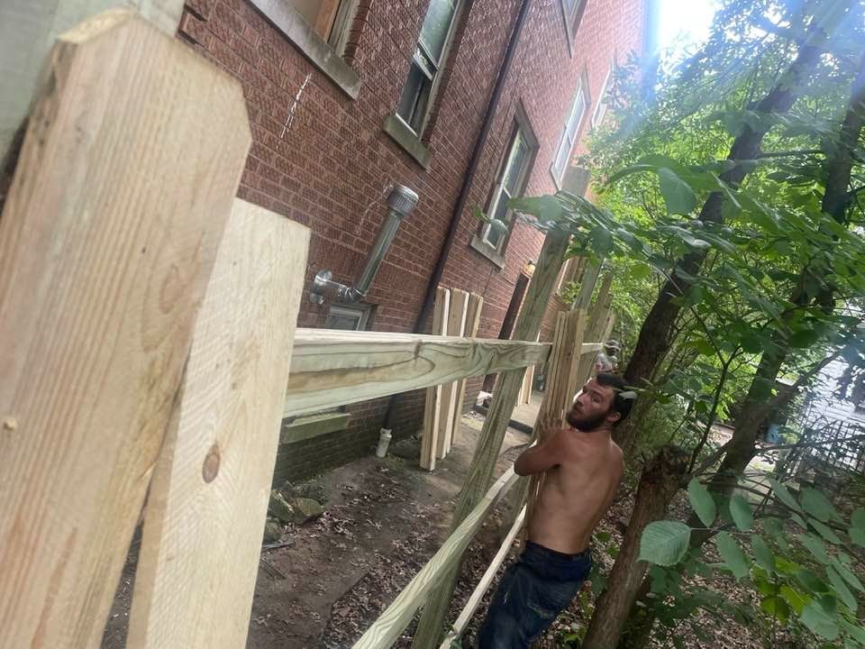 A shirtless person builds a wooden fence next to a brick building.