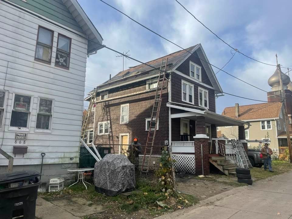 Two-story brown house with ladders, workers. Adjacent white house. Cloudy sky.