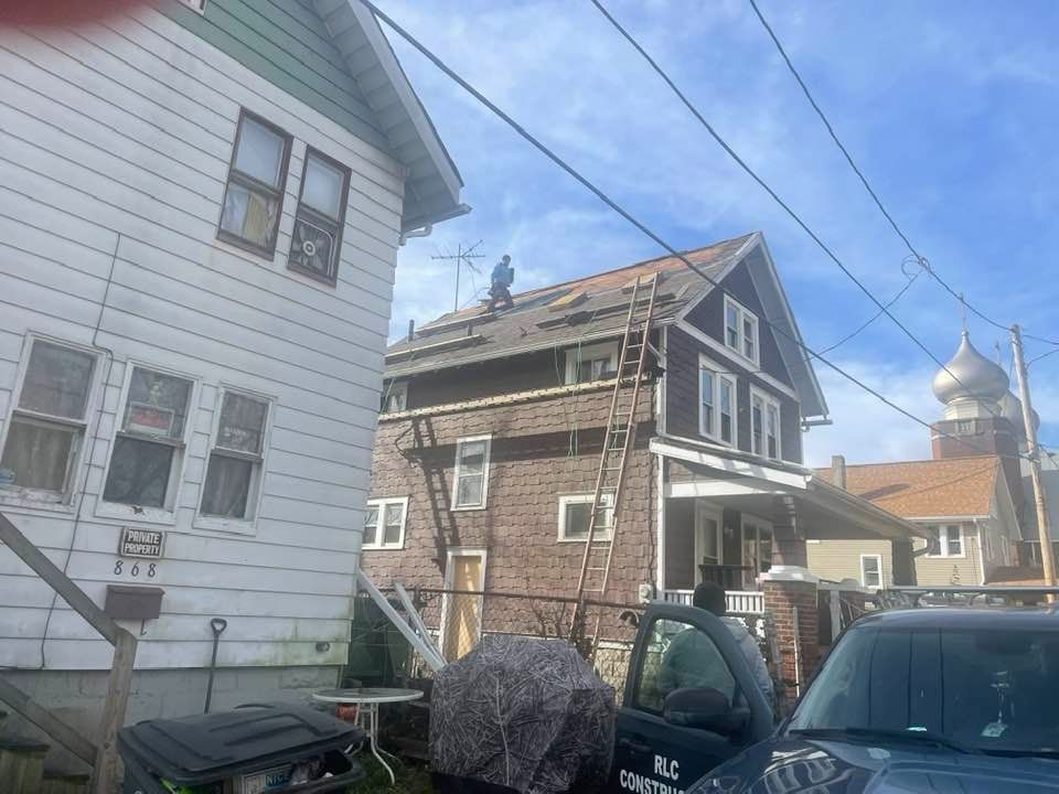 Man on roof of brown house with ladder, wires, and adjacent white house under a blue sky.