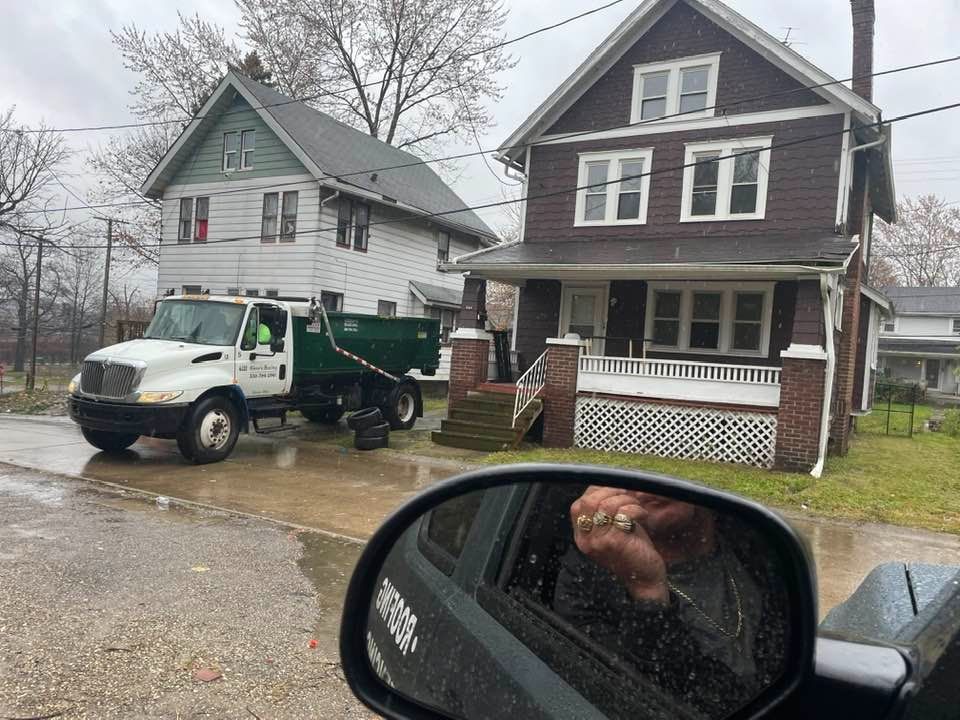 White dump truck parked on a residential street; person in car mirror; two houses in background.