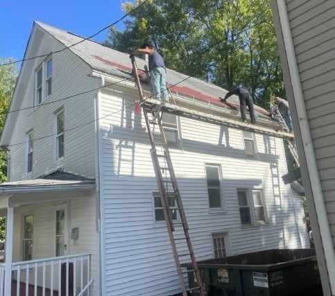 Two workers on a rooftop, replacing shingles. White house, brown ladder, green trees, blue sky, and a dumpster.