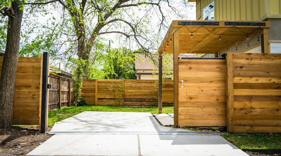 A backyard with a wooden fence, gate, and pergola, concrete patio, and greenery.