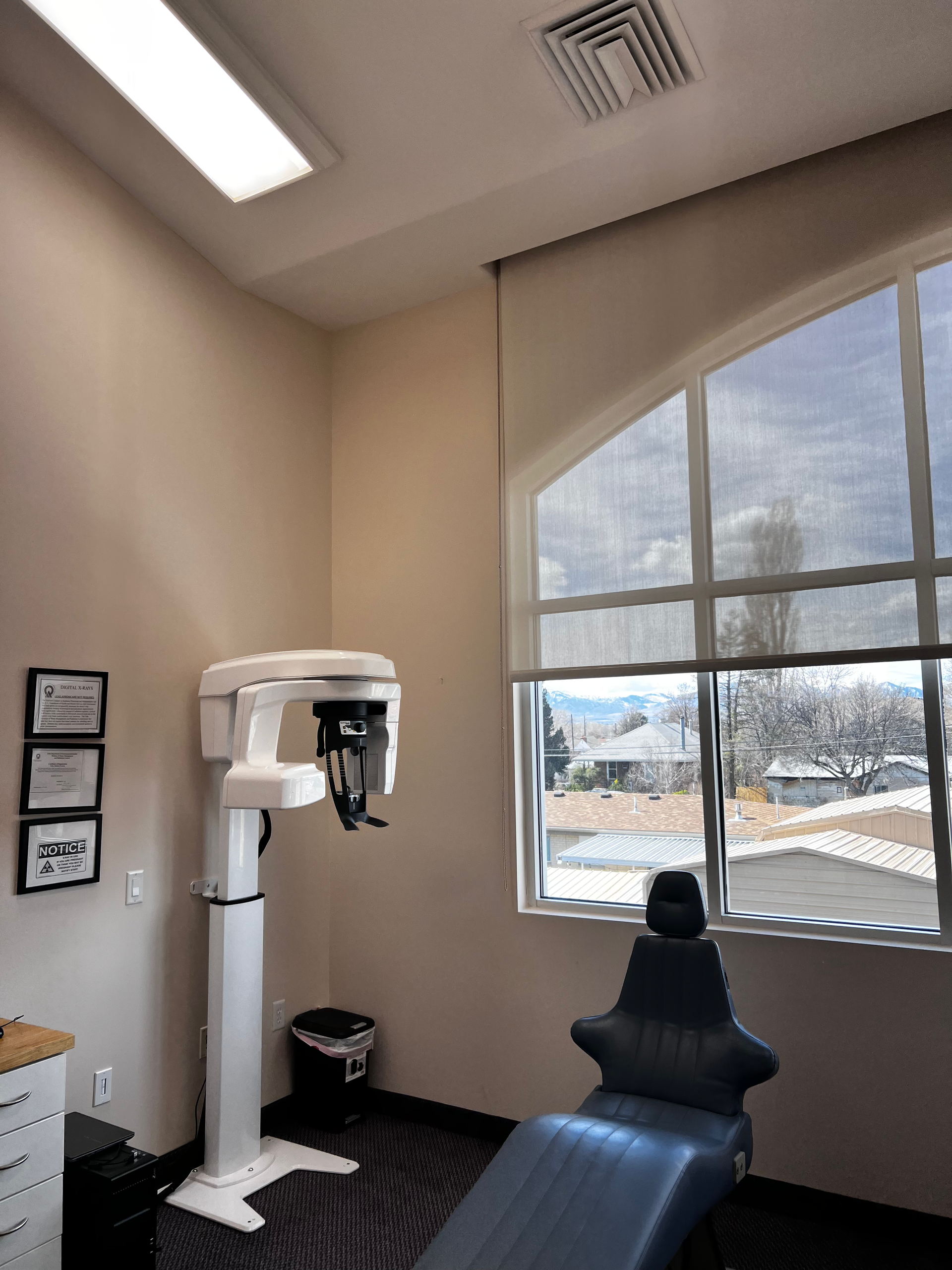 A dental office room with a panoramic X-ray machine standing next to a blue exam chair in front of a window.