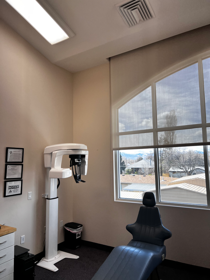 A dental office room with a panoramic X-ray machine standing next to a blue exam chair in front of a window.