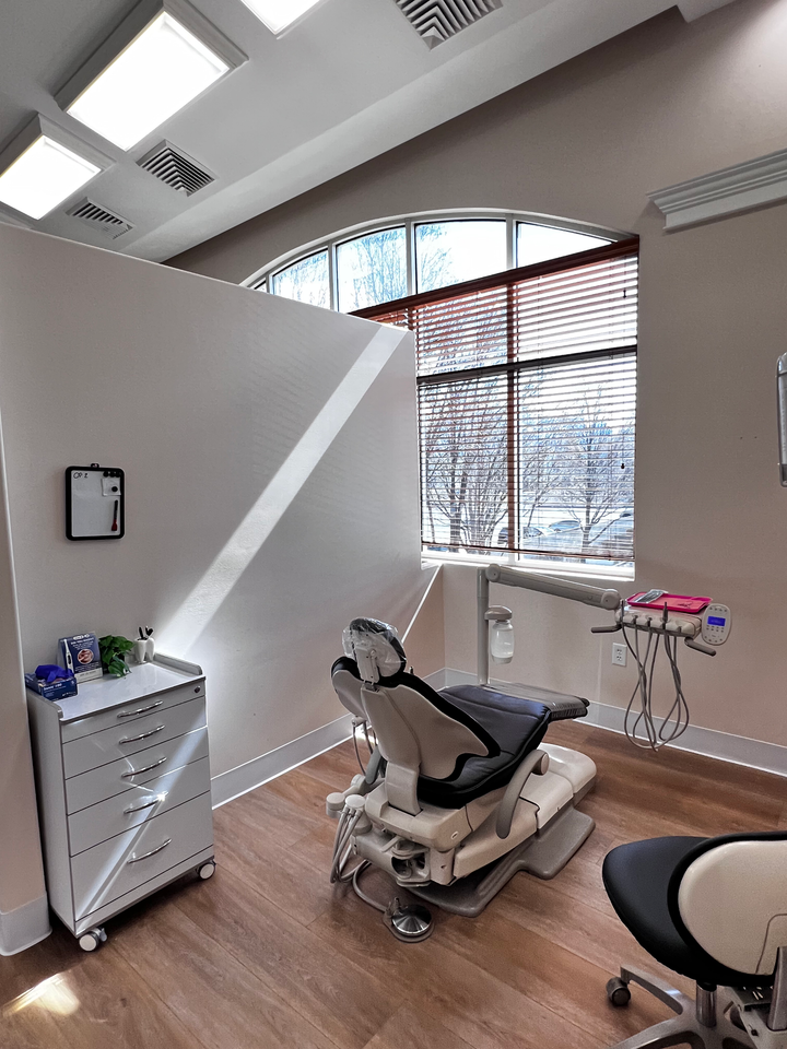 A brightly lit dental exam room with a patient chair, a rolling supply cart, and a large arched window with blinds.