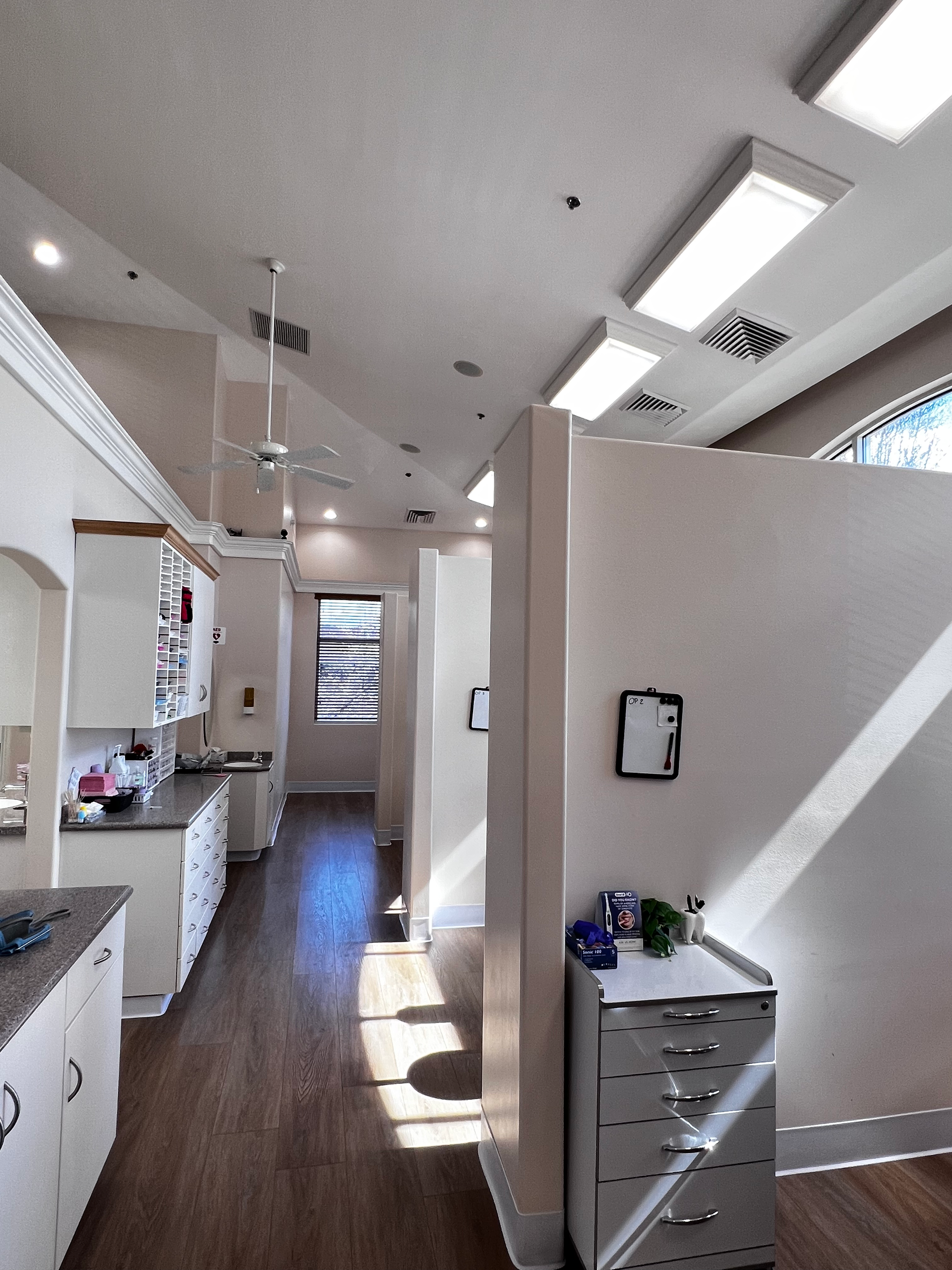 A medical or clinical office with individual partitioned exam areas, white cabinets, wood flooring, and overhead lighting.