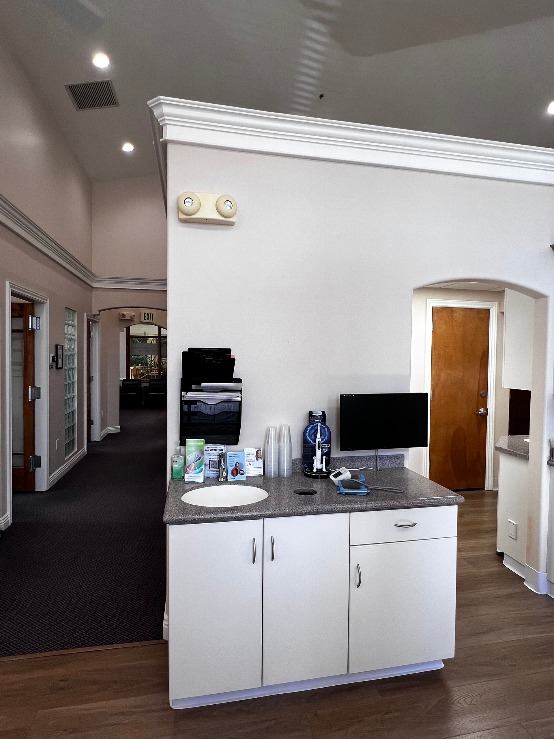 A white cabinet with a small sink and computer monitor sits against a wall in an office hallway with wood-look flooring.