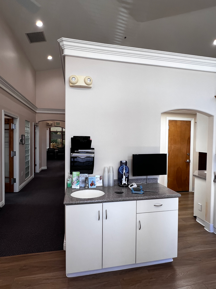 A white cabinet with a small sink and computer monitor sits against a wall in an office hallway with wood-look flooring.
