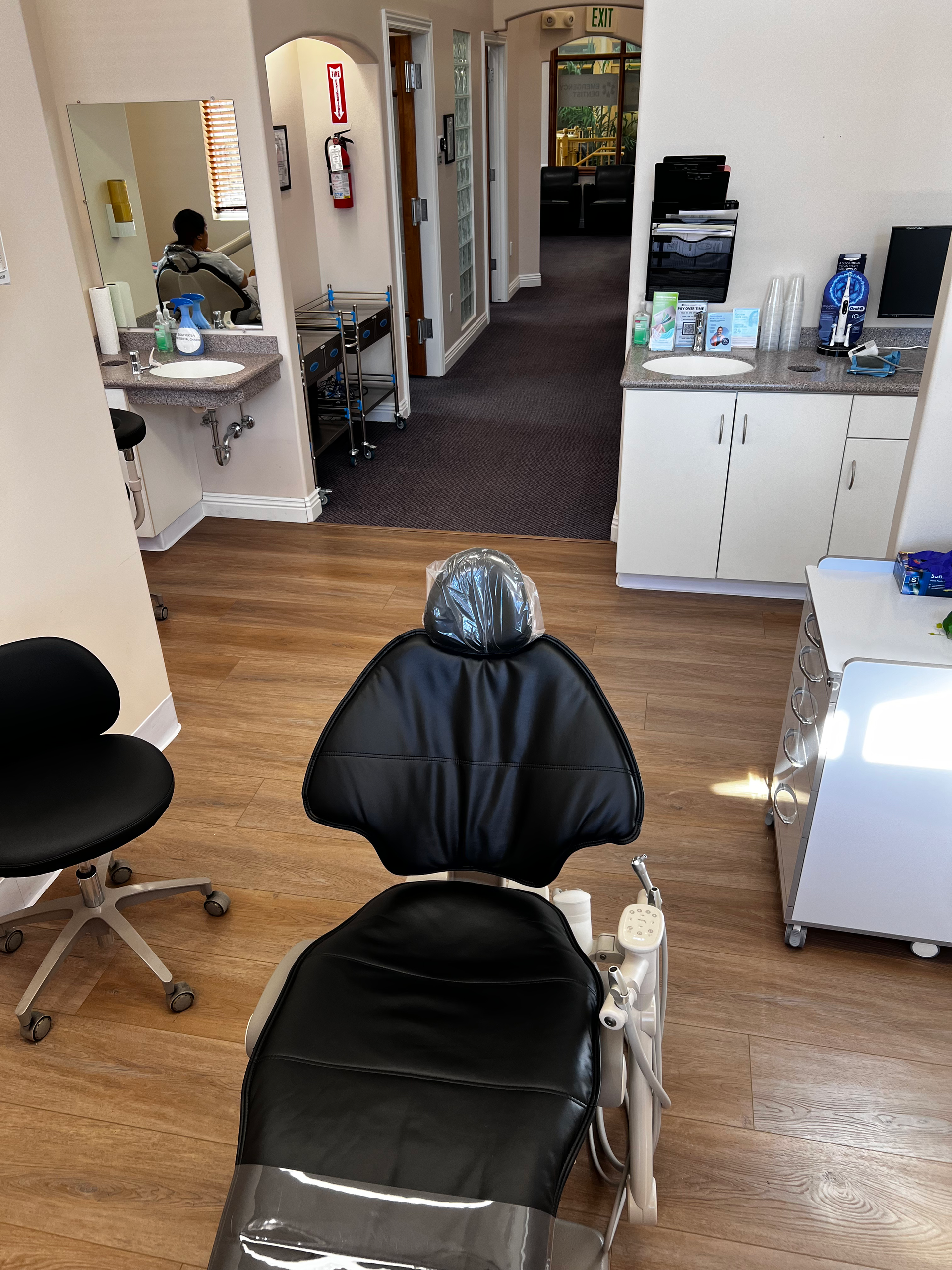 An exam room with a black dental chair, a stool, and white cabinetry. An open hallway is visible in the background.