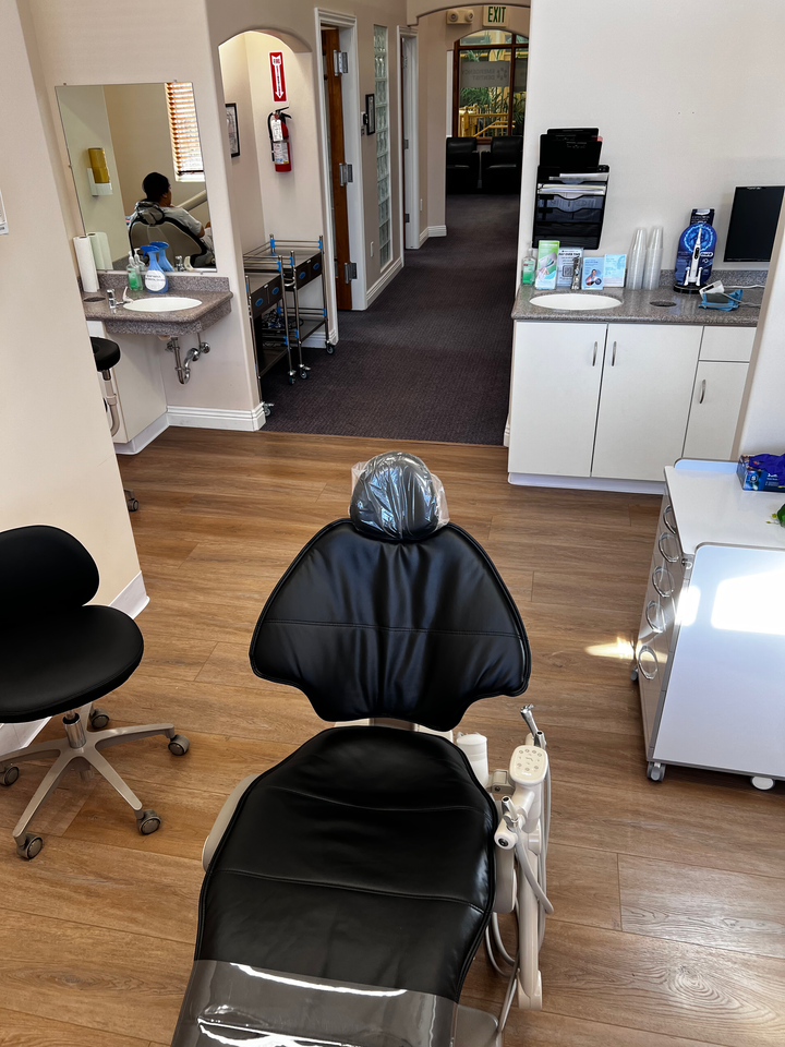 An exam room with a black dental chair, a stool, and white cabinetry. An open hallway is visible in the background.