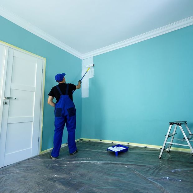 Painter in blue overalls painting a white stripe near the ceiling of a teal-walled room.