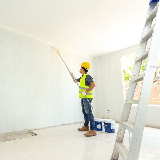Person in safety vest and hard hat painting a white wall with a roller. Ladder and paint buckets visible.