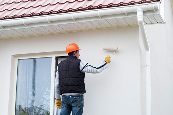 Person in orange hard hat paints the exterior wall of a house with a roller, near a window and gutter.