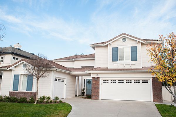 Two-story white house with blue shutters, a two-car garage, and a curved driveway under a blue sky.