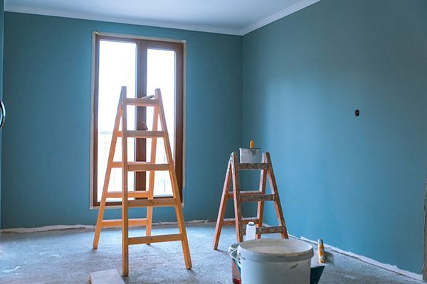 Room being painted blue, with two wooden ladders in front of a window. Paint cans on the floor.